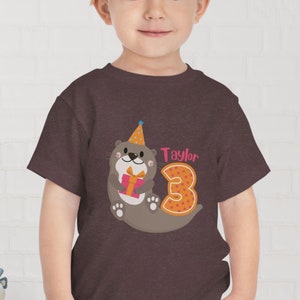 K&ouml;nnte beinhalten: Ein T-Shirt in Heide-Maroon mit einem Motiv einer Cartoon-Otter, die einen Partyhut tr&auml;gt und ein Geschenk h&auml;lt. Die Otter befindet sich neben der Zahl 3. Der Text "Taylor" befindet sich &uuml;ber der Zahl.