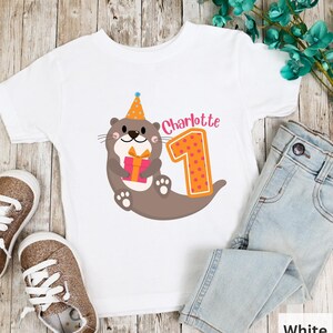 K&ouml;nnte beinhalten: Wei&szlig;es T-Shirt mit einem braunen Otter, der einen Partyhut tr&auml;gt und ein Geschenk h&auml;lt. Der Otter befindet sich neben einer gro&szlig;en orangen Zahl eins mit rosa Punkten. Der Text "Charlotte" befindet sich &uuml;ber der Zahl eins.
