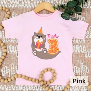 K&ouml;nnte beinhalten: Rosa T-Shirt mit kurzen &Auml;rmeln und einem Aufdruck einer Otter, die einen Partyhut tr&auml;gt und ein Geschenk h&auml;lt. Die Zahl 3 befindet sich neben der Otter und der Name "Taylor" ist &uuml;ber der Otter.