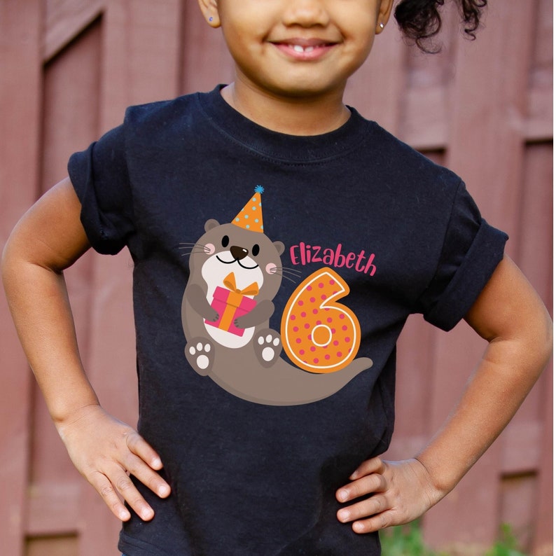 K&ouml;nnte beinhalten: Ein schwarzes T-Shirt mit einer Cartoon-Otter, die einen Partyhut tr&auml;gt und ein Geschenk h&auml;lt. Die Otter befindet sich neben einer gro&szlig;en orangen Zahl sechs mit wei&szlig;en Punkten. Der Text "Elizabeth" ist in Pink &uuml;ber der Zahl gedruckt.