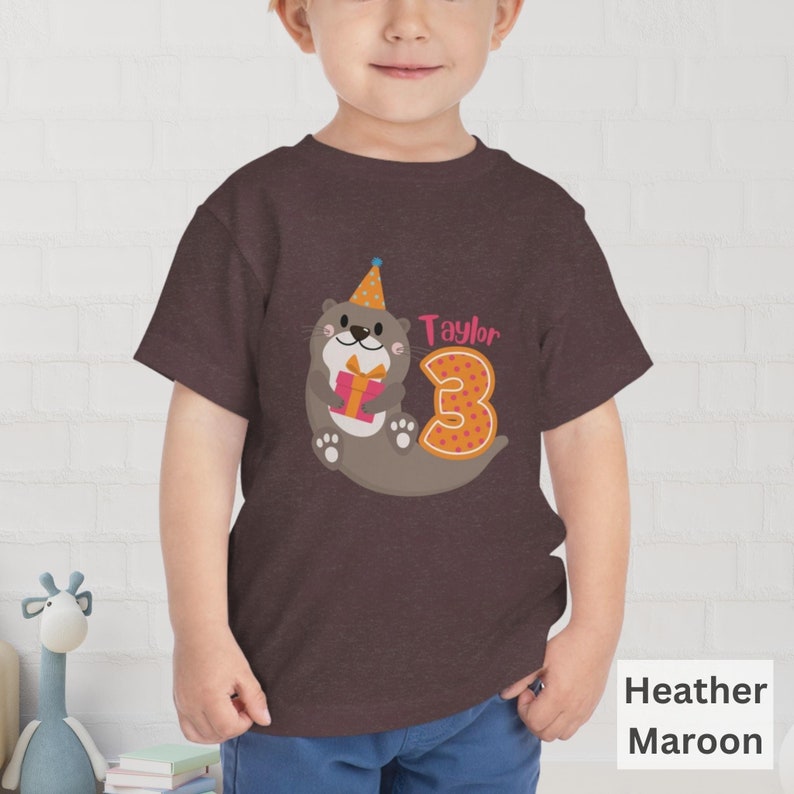 K&ouml;nnte beinhalten: Ein T-Shirt in Heide-Maroon mit einem Motiv einer Cartoon-Otter, die einen Partyhut tr&auml;gt und ein Geschenk h&auml;lt. Die Otter befindet sich neben der Zahl 3. Der Text "Taylor" befindet sich &uuml;ber der Zahl.