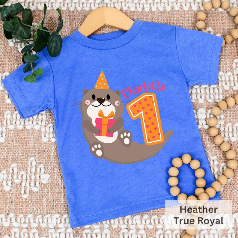 K&ouml;nnte beinhalten: Ein blaues T-Shirt mit einem Cartoon-Otter, der einen Partyhut tr&auml;gt und ein Geschenk h&auml;lt. Der Otter befindet sich neben der Nummer eins mit orangefarbenen Punkten. Der Text "Charlotte" befindet sich &uuml;ber der Nummer eins.