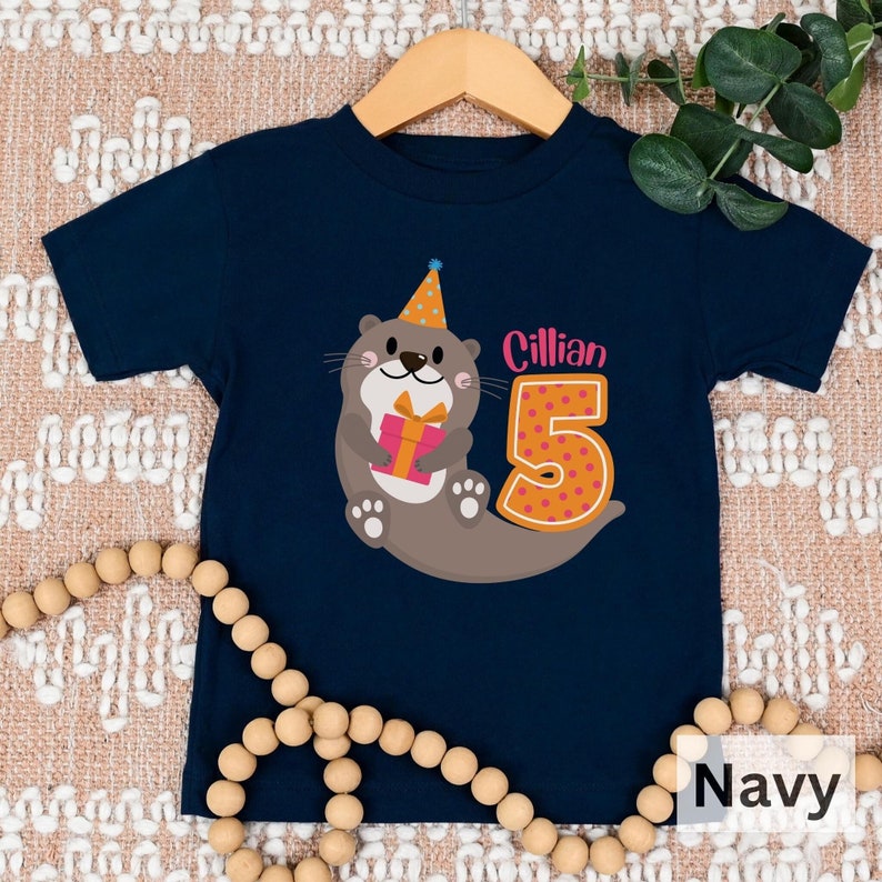 K&ouml;nnte beinhalten: Ein marineblaues T-Shirt mit einem Comic-Otter, der einen Partyhut tr&auml;gt und ein Geschenk h&auml;lt. Das Shirt tr&auml;gt au&szlig;erdem den Namen "Cillian" und die Zahl "5" mit rosa Punkten.