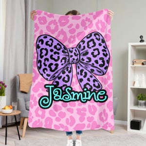 May include: A soft rectangular blanket featuring a pink leopard print background. Centered on the blanket is a large purple bow with a black leopard print pattern. Below the bow, the name Jasmine is written in a bright cyan cursive font.