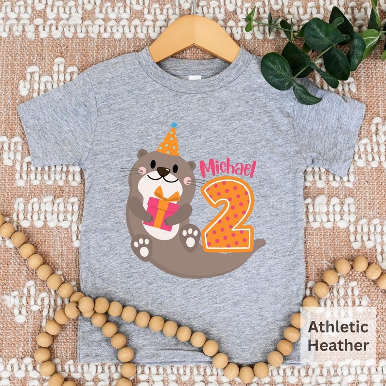 K&ouml;nnte beinhalten: Graues T-Shirt mit kurzen &Auml;rmeln und einem Aufdruck einer Cartoon-Otter, die einen Partyhut tr&auml;gt und ein Geschenk h&auml;lt. Die Zahl 2 befindet sich neben der Otter und hat rosa Punkte. Der Text "Michael" befindet sich &uuml;ber der Zahl. Das Shirt ist aus Athletic Heather Stoff.