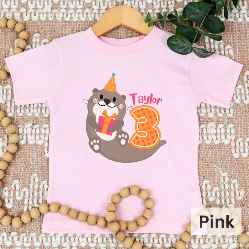 K&ouml;nnte beinhalten: Rosa T-Shirt mit kurzen &Auml;rmeln und einem Aufdruck einer Otter, die einen Partyhut tr&auml;gt und ein Geschenk h&auml;lt. Die Zahl 3 befindet sich neben der Otter und der Name "Taylor" ist &uuml;ber der Otter.