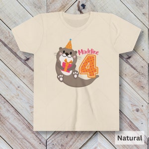 K&ouml;nnte beinhalten: Ein hellbeiges T-Shirt mit einem braunen Otter, der einen Partyhut tr&auml;gt und ein rosa Geschenk h&auml;lt. Der Otter befindet sich neben einer gro&szlig;en orangen Zahl 4 mit rosa Punkten. Der Text "Madeline" befindet sich &uuml;ber der Zahl.