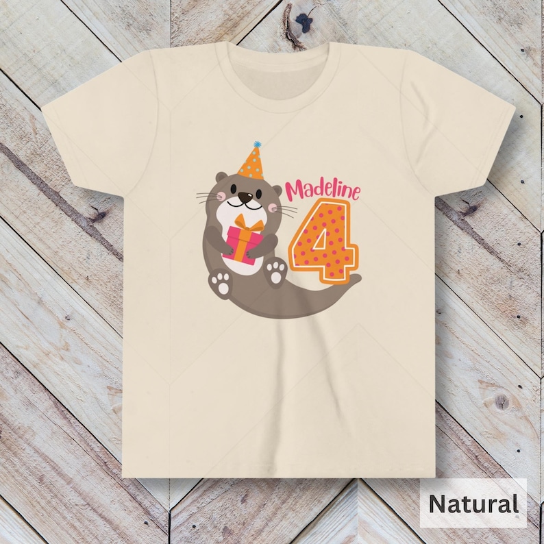 K&ouml;nnte beinhalten: Ein hellbeiges T-Shirt mit einem braunen Otter, der einen Partyhut tr&auml;gt und ein rosa Geschenk h&auml;lt. Der Otter befindet sich neben einer gro&szlig;en orangen Zahl 4 mit rosa Punkten. Der Text "Madeline" befindet sich &uuml;ber der Zahl.