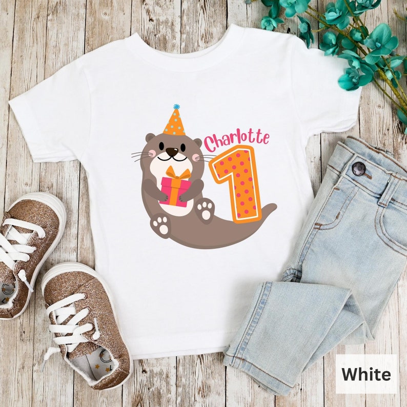 K&ouml;nnte beinhalten: Wei&szlig;es T-Shirt mit einem braunen Otter, der einen Partyhut tr&auml;gt und ein Geschenk h&auml;lt. Der Otter befindet sich neben einer gro&szlig;en orangen Zahl eins mit rosa Punkten. Der Text "Charlotte" befindet sich &uuml;ber der Zahl eins.