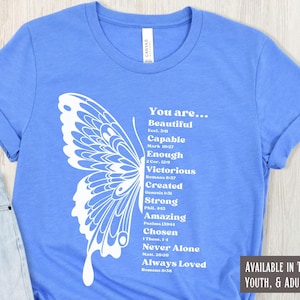 May include: A light blue t-shirt with a white butterfly graphic. The butterfly is made up of words like "Beautiful", "Capable", "Enough", "Victorious", "Created", "Strong", "Amazing", "Chosen", "Never Alone", and "Always Loved". Each word is accompanied by a Bible verse reference.