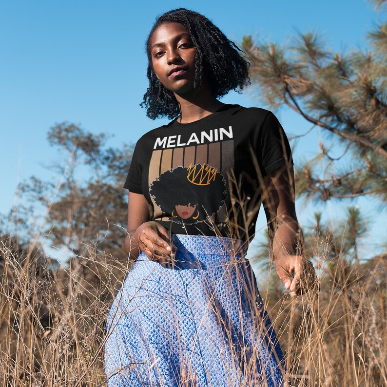 May include: A woman wearing a black t-shirt with the text "MELANIN" and a graphic of a woman with a crown. The graphic is made up of brown and black stripes. The woman is standing in a field of tall grass.