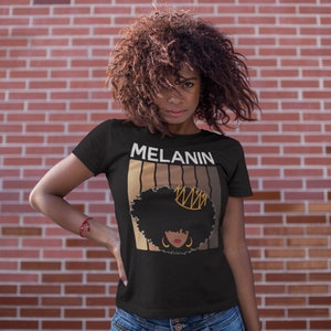 May include: Black t-shirt with the word "MELANIN" printed in white letters. The shirt features a graphic of a woman with an afro hairstyle wearing a gold crown. The graphic is surrounded by a series of brown stripes.