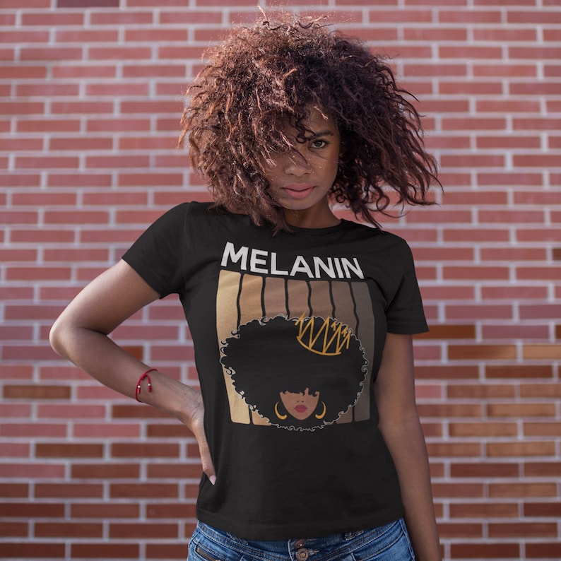 May include: Black t-shirt with the word "MELANIN" printed in white letters. The shirt features a graphic of a woman with an afro hairstyle wearing a gold crown. The graphic is surrounded by a series of brown stripes.
