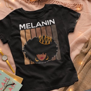 May include: Black t-shirt with the word "MELANIN" in white text and a graphic of a woman with an afro wearing a crown. The graphic is surrounded by brown stripes.