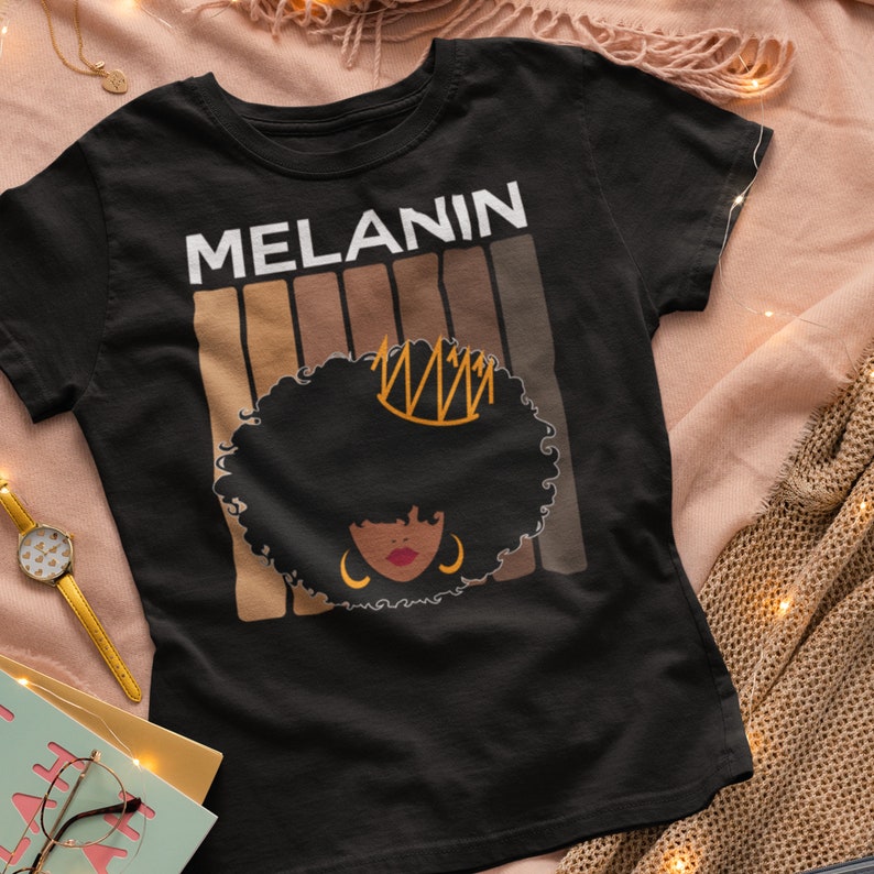 May include: Black t-shirt with the word "MELANIN" in white text and a graphic of a woman with an afro wearing a crown. The graphic is surrounded by brown stripes.