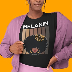 May include: Black t-shirt with the word "MELANIN" in white letters and a graphic of a woman with a crown and afro hair. The graphic is surrounded by a rainbow of brown skin tones.