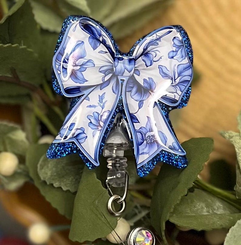 May include: Blue and white floral patterned bow with glitter accents attached to a clear retractable badge reel.