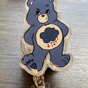 May include: A blue Care Bear with a grumpy expression and a dark blue cloud with raindrops on its belly. The Care Bear is standing with its arms crossed and has a glitter finish. The Care Bear is attached to a clear plastic retractable badge holder.