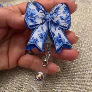 May include: A blue and white floral patterned bow with glitter accents attached to a retractable badge holder with a clear plastic strap and a crystal rhinestone.