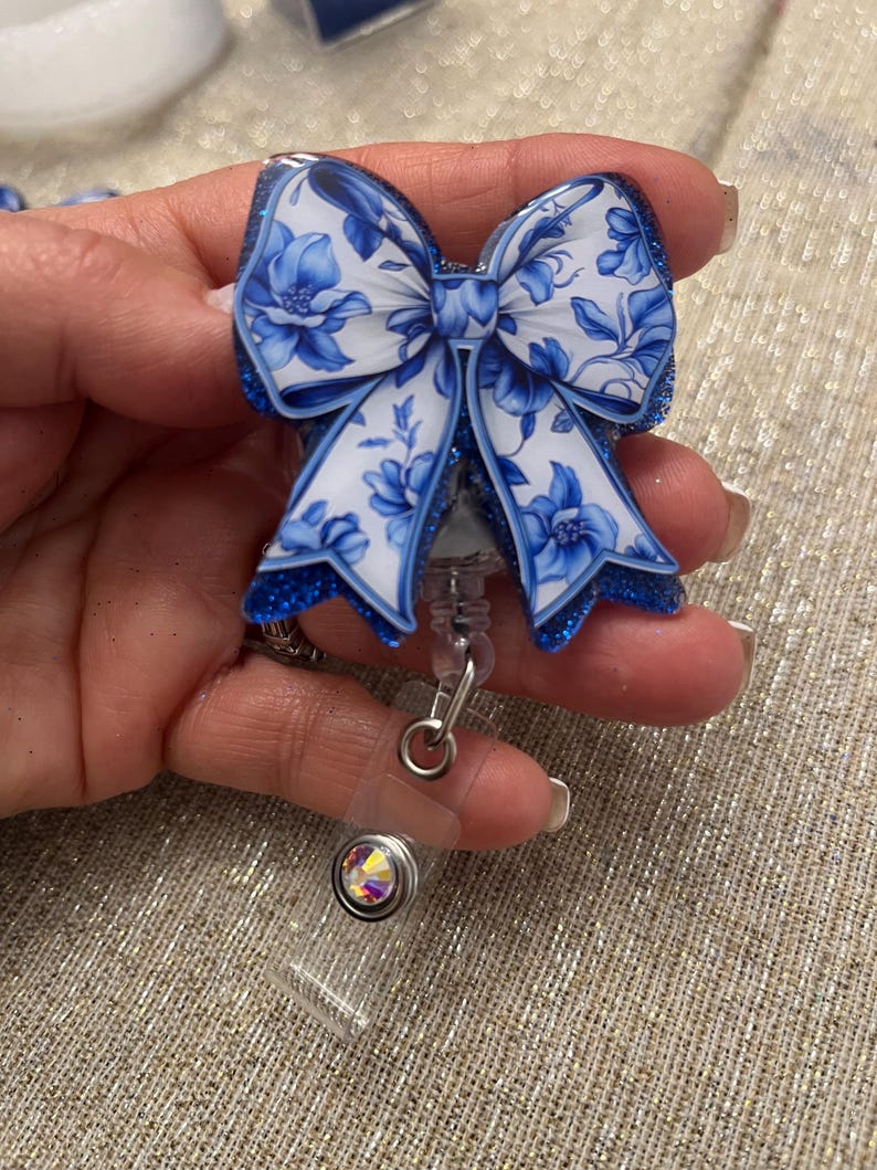 May include: A blue and white floral patterned bow with glitter accents attached to a retractable badge holder with a clear plastic strap and a crystal rhinestone.