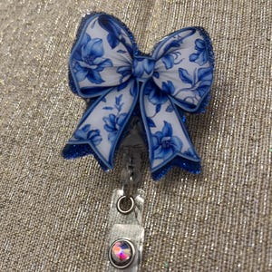 May include: A blue and white floral patterned bow with glitter accents attached to a clear plastic retractable badge holder with a clear rhinestone.