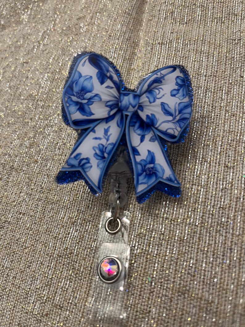 May include: A blue and white floral patterned bow with glitter accents attached to a clear plastic retractable badge holder with a clear rhinestone.