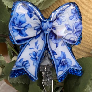 May include: A blue and white floral patterned bow with glitter accents attached to a clear retractable badge holder with a silver ring.