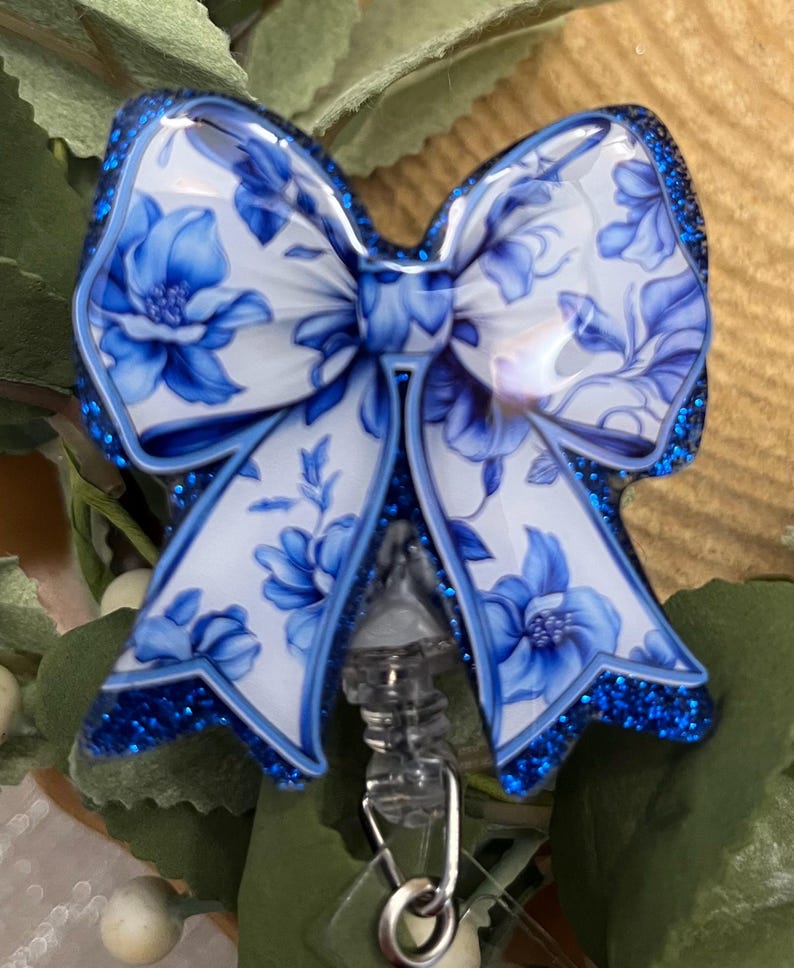 May include: A blue and white floral patterned bow with glitter accents attached to a clear retractable badge holder with a silver ring.