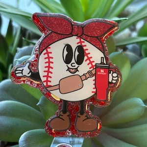 May include: A baseball-themed badge reel featuring a cartoon baseball character with a red bow, holding a red Stanley tumbler. The character has brown shoes and a brown belt. The badge reel has a red glitter border.