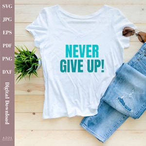 May include: White t-shirt with the text "NEVER GIVE UP!" in teal blue lettering. The shirt is laid flat on a wooden surface with a pair of blue jeans and sunglasses.