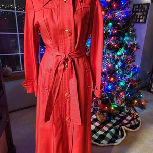 May include: A long red coat with a belt and gold buttons. The coat has a collar and long sleeves. The coat is photographed in front of a Christmas tree.