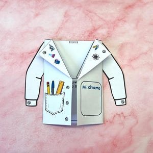 Può includere: Un camice da laboratorio di carta bianca con contorni neri, con una tasca con penne e un righello, e adesivi decorativi a tema scientifico. La frase italiana "Mi chiamo" è scritta sul lato destro. Sfondo rosa marmorizzato.