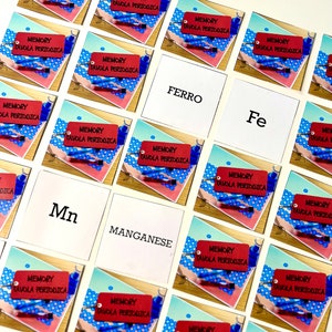Può includere: Un gioco di memoria con carte rosse e bianche che mostrano la tavola periodica degli elementi. Le carte hanno le parole "Memory Tavola Periodica" stampate su di esse. Le carte sono disposte in una griglia su una superficie bianca. Le parole "Ferro", "Fe", "Mn" e "Manganese" sono stampate su carte bianche.