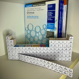 May include: A white periodic table of elements model with a black and white label that reads "Tabella periodica degli elementi". The model is made of cardboard and is sitting on a wooden shelf. The model is in a classroom setting.