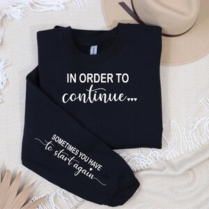 May include: Black sweatshirt with white text that reads "In order to continue... Sometimes you have to start again".