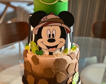 Può includere: Una torta a tre piani decorata con un personaggio di Topolino che indossa un cappello da safari. La torta è decorata con foglie verdi, strisce bianche e marroni e piastrelle esagonali marroni. Il livello inferiore ha il nome "Benjamin" scritto in lettere dorate.
