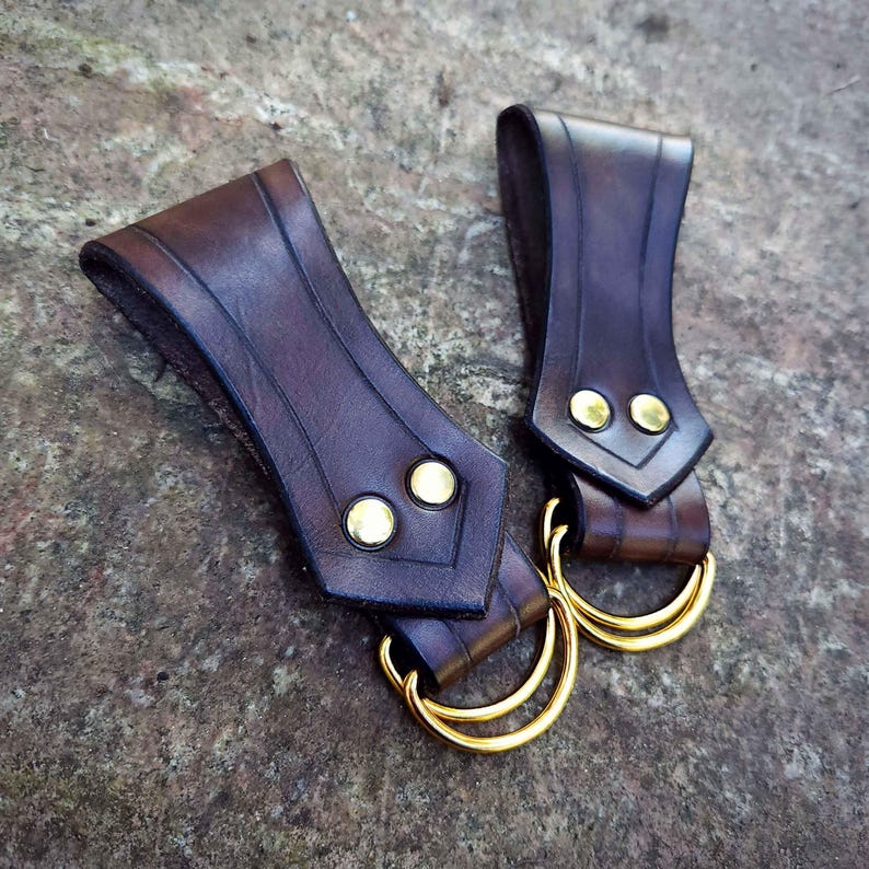 May include: Two brown leather sword belt loops with gold metal rings. The loops have a decorative design and are secured with gold rivets.