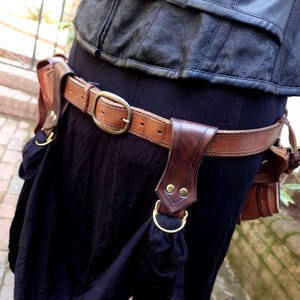 May include: A brown leather belt with a large brass buckle and two leather pouches attached. The belt is worn over a black skirt.