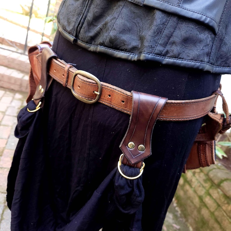May include: A brown leather belt with a large brass buckle and two leather pouches attached. The belt is worn over a black skirt.