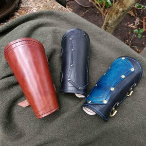May include: Three leather arm guards in reddish-brown, black, and blue. The arm guards are cylindrical with decorative stitching and metal accents. The blue arm guard has a lighter blue section.