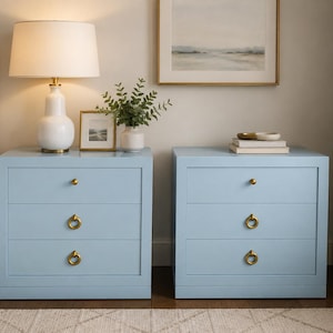 May include: Two light blue wooden nightstands, each featuring three drawers with brass hardware. The top drawer has a small round knob, while the two lower drawers are equipped with circular ring pulls. The cabinets have a smooth, matte finish.