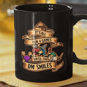 Könnte beinhalten: Schwarze Keramik-Tasse mit dem Text "It's all fun & games until the DM smiles" in einem Banner-Design. Die Tasse zeigt Cartoon-Illustrationen von Fantasy-Spielcharakteren und -elementen. Die Tasse steht auf einer gelben Oberfläche.