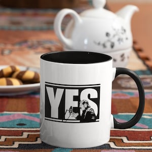 May include: White ceramic mug with a black rim and handle. The mug has a black and white graphic of a person holding a microphone with the word "YES" in large letters.