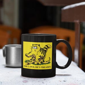May include: Black coffee mug with a yellow background featuring a cartoon cat stepping on a snake. The text on the mug reads "OH LAWD, HE'S TREADIN"
