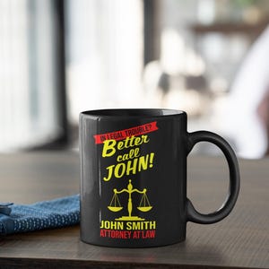 May include: Black coffee mug with a yellow and red design. The mug says "In legal trouble? Better call John!" and "John Smith Attorney at Law".