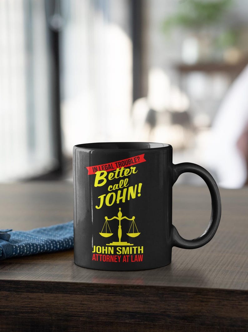 May include: Black coffee mug with a yellow and red design. The mug says "In legal trouble? Better call John!" and "John Smith Attorney at Law".
