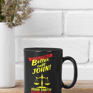 May include: Black coffee mug with a white background and the text "In legal trouble? Better call JOHN!" in yellow and red. Below the text is a yellow scale of justice and the text "JOHN SMITH ATTORNEY AT LAW" in white.