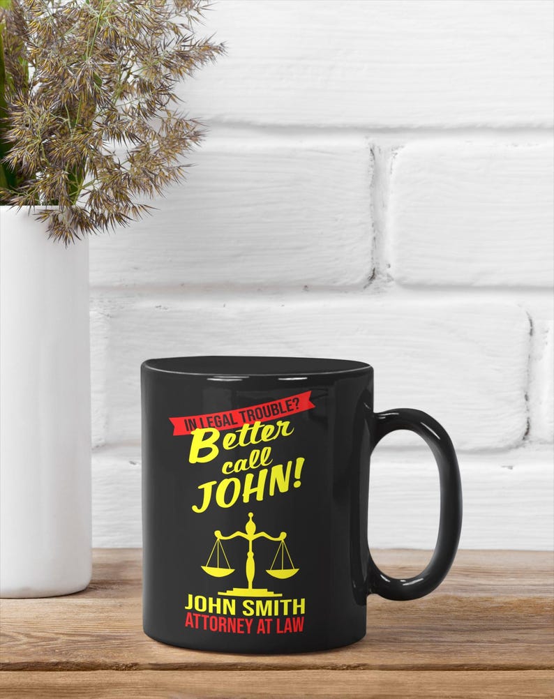 May include: Black coffee mug with a white background and the text "In legal trouble? Better call JOHN!" in yellow and red. Below the text is a yellow scale of justice and the text "JOHN SMITH ATTORNEY AT LAW" in white.