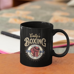 May include: Black coffee mug with the text "Cutty's Boxing" in a vintage style. The design includes a boxing glove and the words "Boxing Gym, West Side, Baltimore, Maryland."