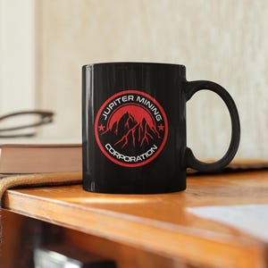 Può includere: Tazza da caffè nera con un logo rosso e bianco che dice "Jupiter Mining Corporation" con una grafica di catena montuosa all'interno del logo.